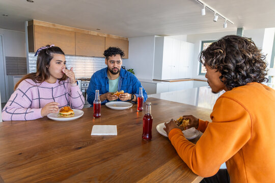 Diverse friends sharing hamburgers and sipping red soda at wooden kitchen island in casual clothing