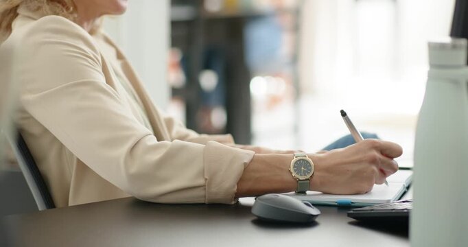 Woman in 30s writing in notebook at desk during planning, pausing, holding pen to chin