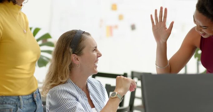 Diverse female coworkers celebrating win high-fiving at desk, seated woman raising hand by monitor