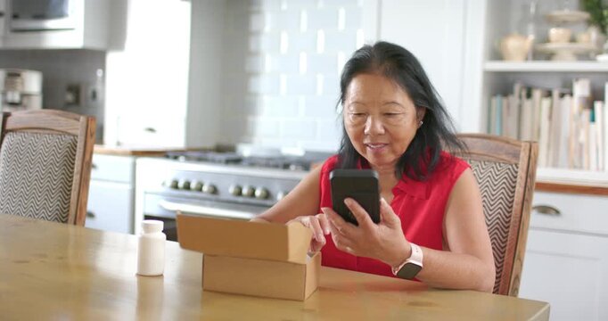Senior Asian woman in red top opening package at kitchen checking smartphone and inspecting bottle