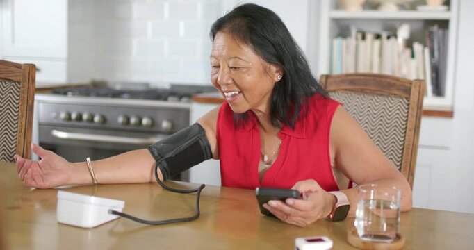 Senior Asian woman wearing red blouse sitting at kitchen table, tapping phone, monitoring BP cuff