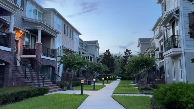 A row of contemporary apartment buildings featuring light blue and tan siding is illuminated by warm bollard lights along a concrete path. The manicured green lawn and mature trees create a peaceful
