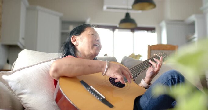 Senior Asian woman settling on couch strumming acoustic guitar and singing for rest at home