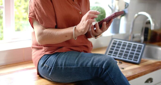 Senior Asian woman rust top on bench tapping phone seeing cable plugging into solar panel charging