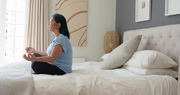 Senior Asian woman sitting on bed near window after settling, holding mudra and breathing for calm