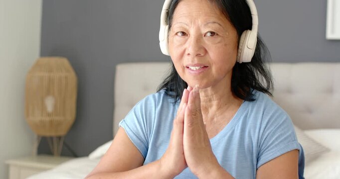 Senior Asian woman holding palms together on bed, hearing audio on white headphones, smiling