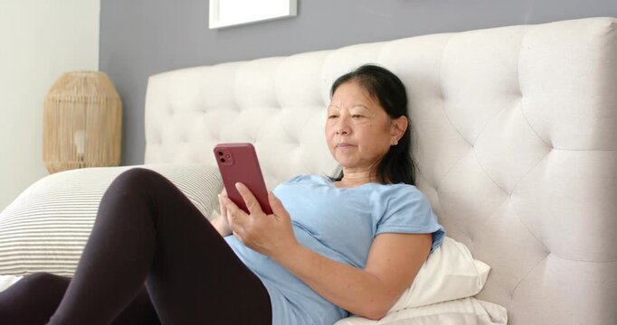 Senior Asian woman in blue tee holding maroon phone tapping after alert smiling by tufted headboard