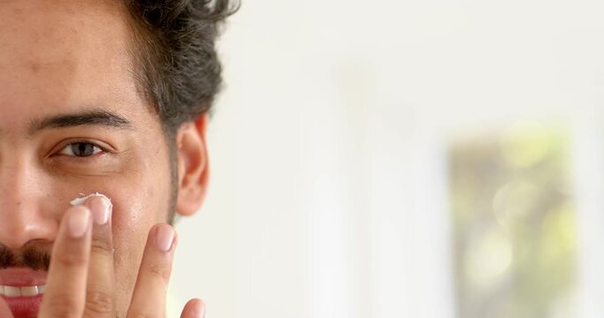Man in 30s starting with cream dab, smiling, spreading moisturizer on cheek at vanity, copy space