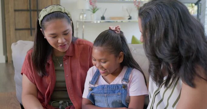 Diverse women carers sharing smiles, adjusting overalls strap, tickling child on couch for bonding