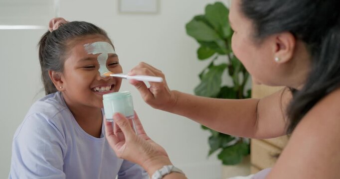 Mother and daughter on bed, mother dipping brush into jar then applying mint mask for pampering