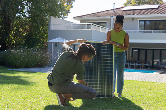 African American couple kneeling and standing, positioning solar panel on lawn by pool and deck
