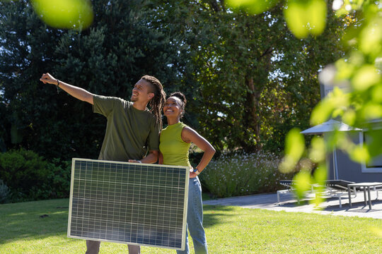 African American couple holding solar panel, pointing outward at home, woman in lime top, jeans