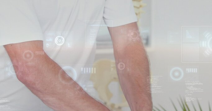 Leaning male torso and forearms wearing white polo over table in clinic with HUD, spine, plant