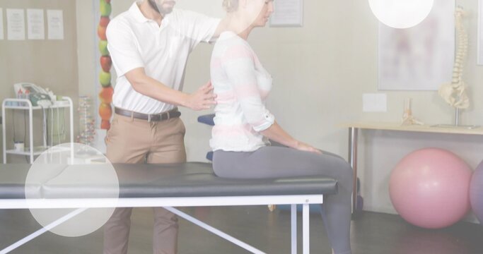 Male clinician in white polo guiding seated female patient in therapy room, with spine model