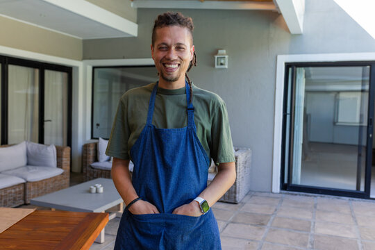 African American man standing and smiling on veranda wearing denim apron, hands tucked in pocket