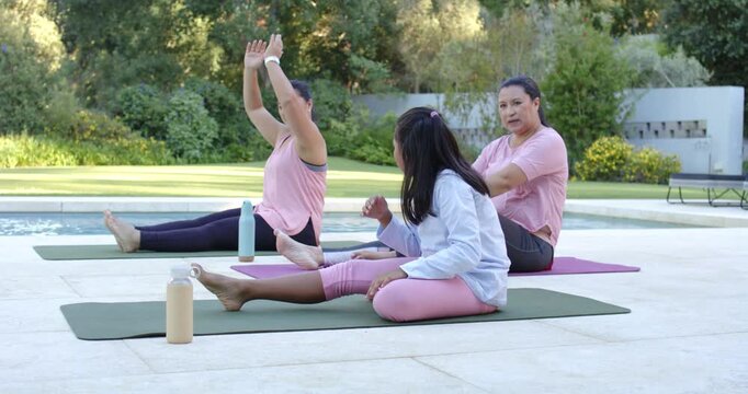 Diverse female family following midlife woman raising arms, reaching toes on mats by pool, bottles
