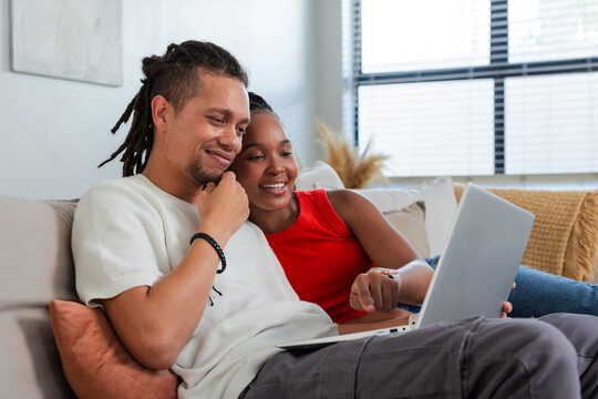 African American couple sitting on sofa leaning over laptop, woman in red sleeveless top pointing