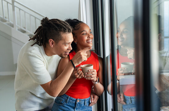 African American couple standing by glass door, man pointing outside, woman in red holding mug