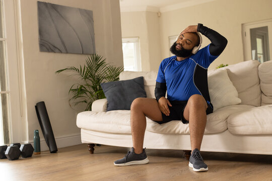 African American adult male sitting cooling down on cream sofa with dumbbells, headphones, water