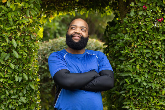 African man standing with arms crossed, looking upward in hedge arch wearing blue shirt