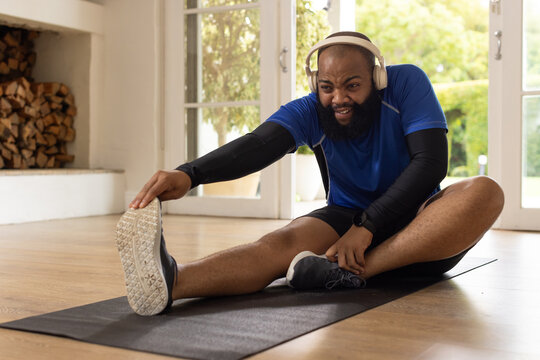 African American man stretching on black mat at home wearing blue shirt, headphones, smartwatch