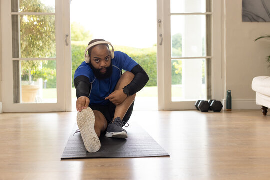 African man stretching hamstring on black mat at home, wearing white headphones, dumbbells visible
