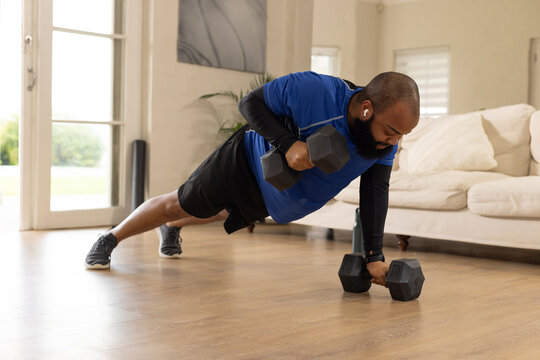 African American man doing renegade row at home wearing workout gear with hex dumbbells and earbuds