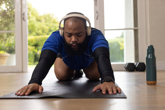African man stretching forward on mat in home wearing blue shirt, beige headphones, wristwatch