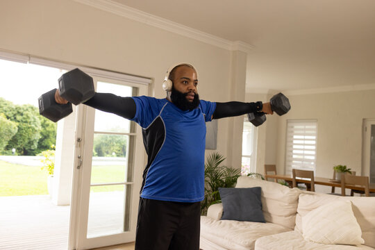 African American man doing lateral raises at home holding hex dumbbells wearing blue athletic shirt
