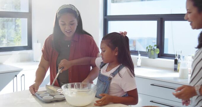 Diverse family helping child ladling batter into muffin tray from mixing bowl on counter, baking