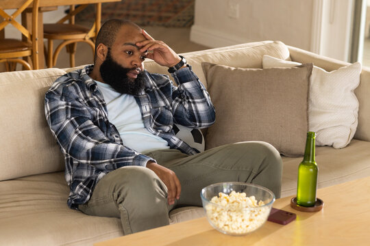 African American man sitting on beige sofa in living room, touching forehead, popcorn bowl nearby