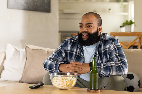 African American man sitting on sofa wearing flannel, grabbing popcorn and remote in living room