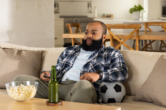 African man sitting on sofa in living room holding TV remote and soccer ball beside