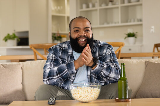 African American man in plaid flannel shirt clapping while watching TV in living room with popcorn