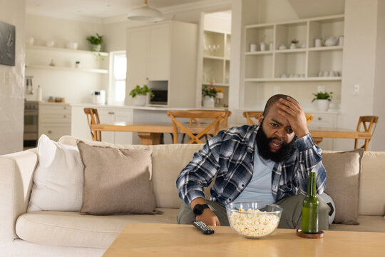 Bearded man wearing plaid overshirt sitting on right side of sofa holding remote near popcorn bowl