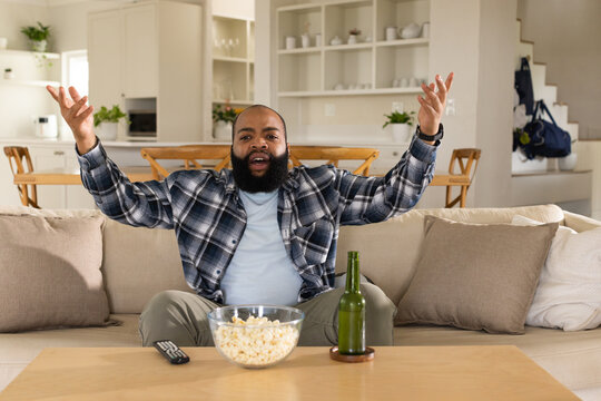 African American man wearing plaid shirt on beige sofa at home watching TV with popcorn, beer