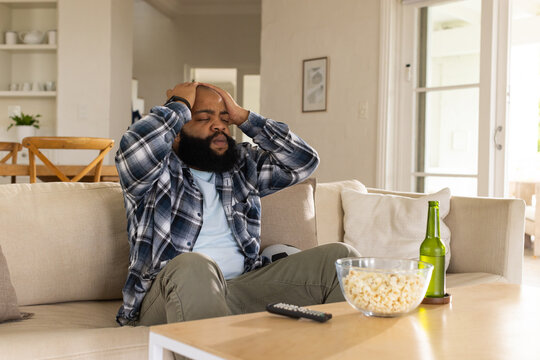 African American man sitting on beige sofa at home, holding head, plaid shirt, popcorn on table