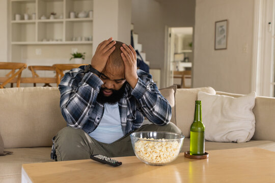 Adult man wearing plaid shirt sitting in living room on beige sofa holding head, popcorn bowl