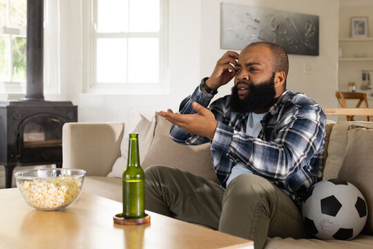 African man wearing plaid shirt sitting on beige sofa in living room, gesturing at popcorn