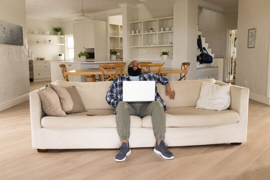 African American man in flannel sitting on sofa at home with two throw pillows using laptop