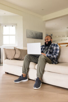 African American man sitting on beige sofa at home smiling at laptop in blue flannel shirt