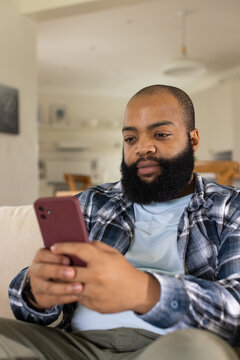 African American man sitting on light sofa at home holding maroon phone, wearing plaid shirt