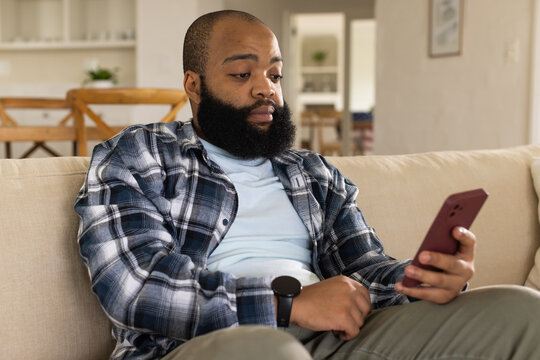 African American man sitting on sofa at home in plaid shirt using maroon phone, black smartwatch
