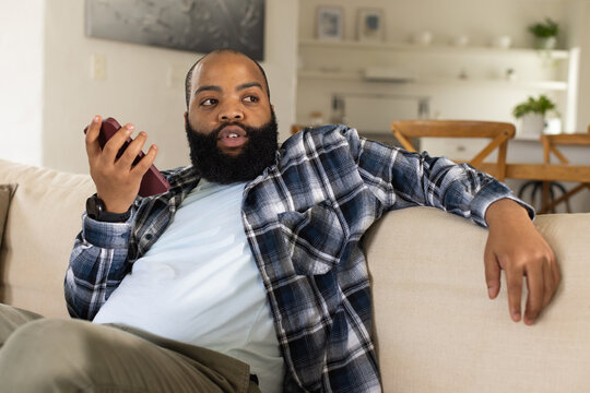African American man reclining on beige sofa in living room, holding maroon smartphone near mouth