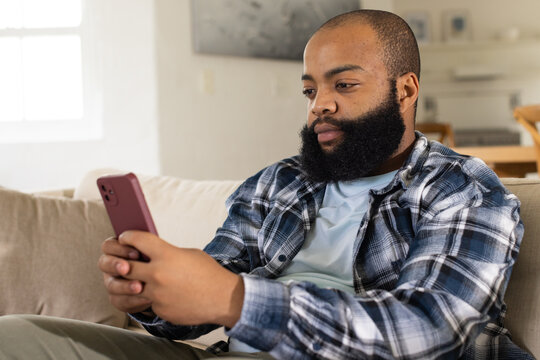 African American man sitting on beige sofa using red smartphone, looking at screen, copy space