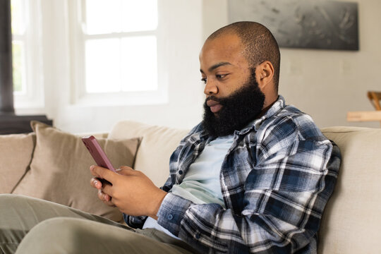 African man sitting on sofa in living room, plaid shirt olive pants, holding pink phone