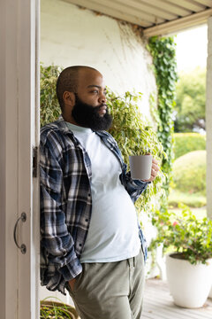 Adult African man wearing plaid shirt leaning in doorway, holding ceramic mug, gazing toward garden