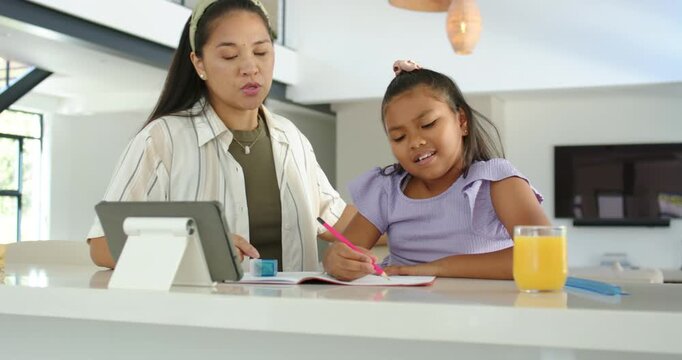 Diverse mother guiding school-age daughter using tablet for homework, writing in notebook at home