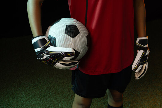 African American woman goalie standing on turf at night, wearing red kit and gloves, holding ball