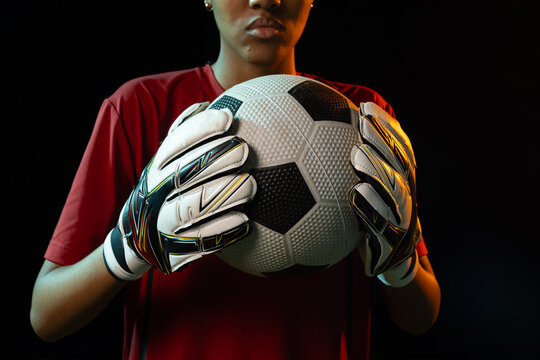 African American female athlete holding soccer ball with gloved hands in studio with red jersey